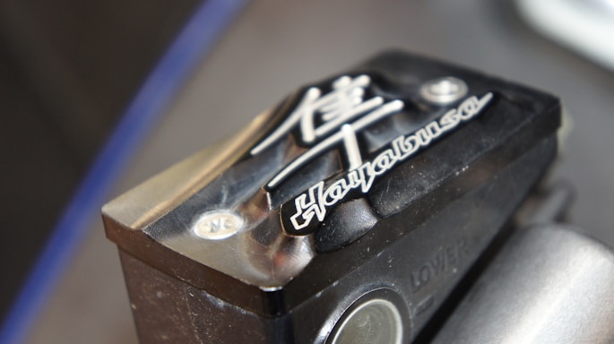 A close-up of a motorcycle part with embossed text and symbols. The text 'Hayabusa' is prominently featured alongside a stylized Japanese character. The part has a metallic, angular design with a black finish and highlights visible on its surface.
