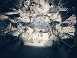 A kaleidoscopic view of an outdoor scene is framed by escalators in a building entrance or exit, with reflections of people and urban surroundings in angular, mirrored surfaces. Pedestrians cross a street in a bustling area filled with buildings and cars in the background, creating a dynamic and busy environment.