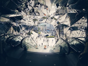 A kaleidoscopic view of an outdoor scene is framed by escalators in a building entrance or exit, with reflections of people and urban surroundings in angular, mirrored surfaces. Pedestrians cross a street in a bustling area filled with buildings and cars in the background, creating a dynamic and busy environment.