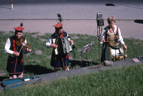 Three musicians in traditional attire perform outdoors. One plays a violin, another an accordion, and the third musician plays a drum. They are standing on grass with a pavement in the background.
