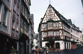 A picturesque street featuring traditional, ornately designed half-timbered buildings. The facades are adorned with balconies and flower boxes, creating a charming and historic atmosphere. People are walking along the cobblestone pavement, adding life to the scene under a cloudy sky.