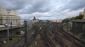 The image depicts a wide view of a railway station with numerous train tracks converging towards the center. There are large, historic-looking buildings flanking the tracks on either side. The sky is overcast with heavy grey clouds, giving a dramatic backdrop to the scene.