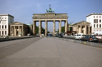 The image features the iconic Brandenburg Gate, a neoclassical monument in Berlin, Germany. It is seen from the front, with its massive pillars and quadriga sculpture on top. Cars and pedestrians are present, suggesting a busy urban setting.