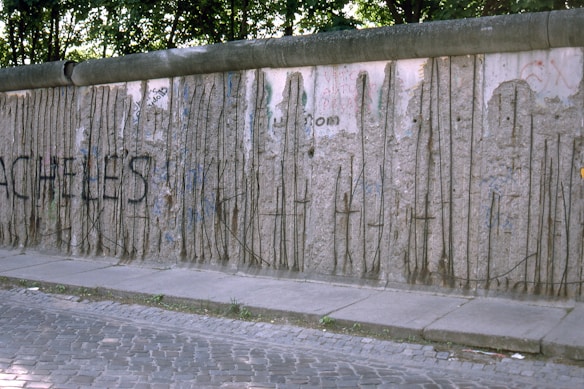 A section of a concrete wall with visible rebar and graffiti. The wall appears weathered with peeling and crumbling areas. Surrounding the wall are green trees casting shadows, and a cobblestone pathway runs alongside it.