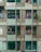 A weathered apartment building facade with visible balconies where laundry is hanging out to dry. The structure has a mix of brickwork and patterned concrete blocks, and the paint is faded. Electrical wires run along the building, and some greenery is visible in planters.