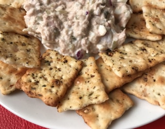 A plate filled with seasoned crackers arranged around a generous serving of a creamy tuna salad mixture. The tuna salad contains visible chunks of vegetables and possibly herbs.