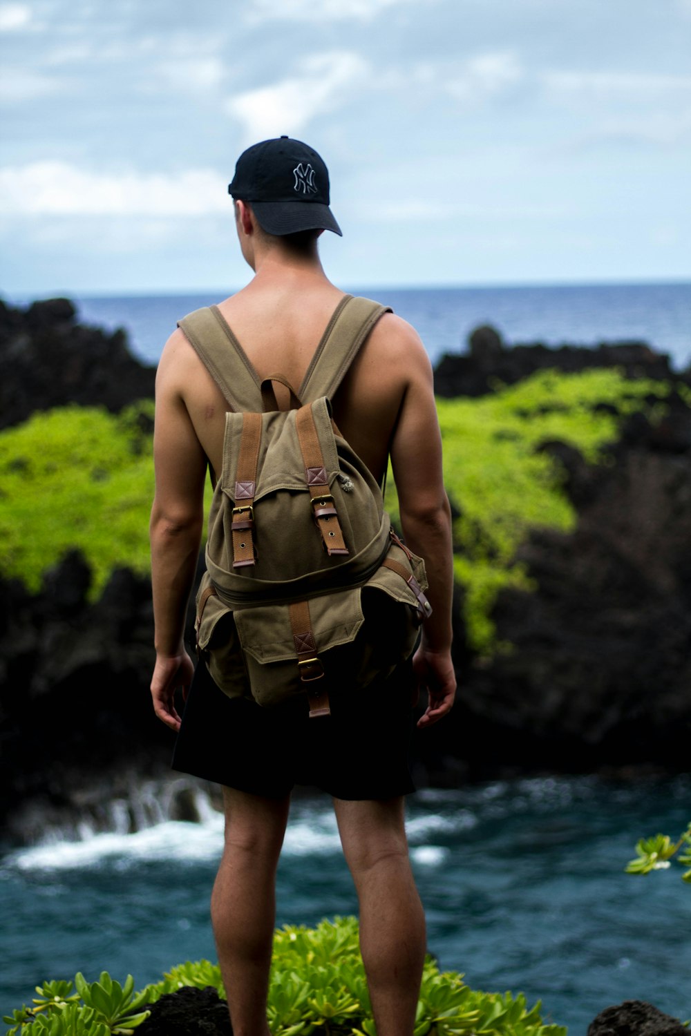 homme debout sur la falaise portant un short noir, une casquette ajustée noire des Yankees de New York et un sac à dos marron pendant la journée