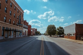 A quiet, empty urban street with a historic building labeled 'Hotel Missouri' on the left. The buildings are mostly made of brick, and the road stretches into the distance. Trees line the street, and the sky is clear with some fluffy clouds.
