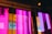 Vibrant vertical neon lights in shades of pink and purple create an abstract, modern aesthetic. The colorful reflections and blurred background contribute to an urban, futuristic vibe.