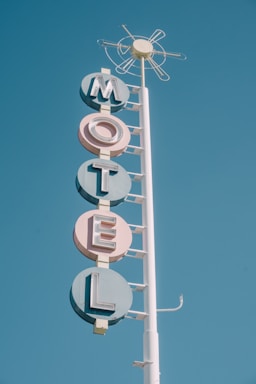 A vintage motel sign with bold, pastel-colored letters spelling out 'MOTEL' stacked vertically. The sign is mounted on a tall white pole against a clear blue sky. The design features retro geometric shapes and a circular element at the top resembling an abstract sun.