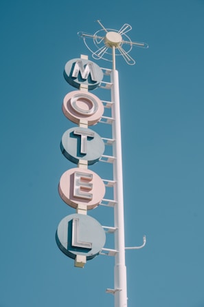A vintage motel sign with bold, pastel-colored letters spelling out 'MOTEL' stacked vertically. The sign is mounted on a tall white pole against a clear blue sky. The design features retro geometric shapes and a circular element at the top resembling an abstract sun.