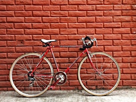 A red road bike with drop handlebars is positioned against a textured brick wall. The bike features thin tires and a white saddle, and there is a clear view of its chain and pedal system. The brick wall is painted a muted orange-red, adding a rustic, urban backdrop.