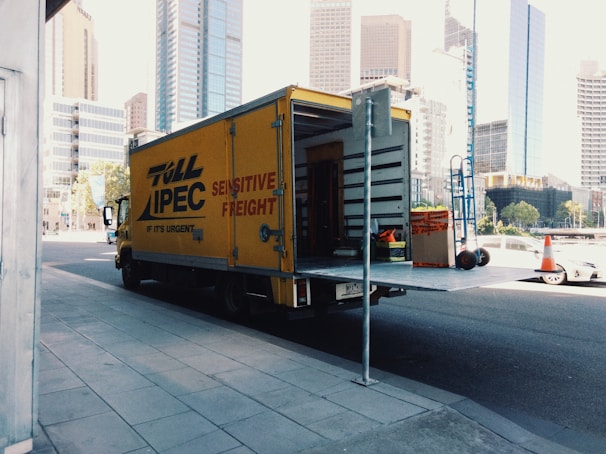 A yellow delivery truck parked on a city street with its rear door open, revealing some equipment and boxes inside. The truck has 'Toll IPEC Sensitive Freight' written on its side. There are tall modern buildings in the background, and a few traffic cones visible near the sidewalk.