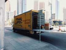 A yellow delivery truck parked on a city street with its rear door open, revealing some equipment and boxes inside. The truck has 'Toll IPEC Sensitive Freight' written on its side. There are tall modern buildings in the background, and a few traffic cones visible near the sidewalk.