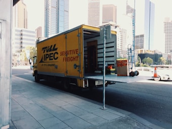A yellow delivery truck parked on a city street with its rear door open, revealing some equipment and boxes inside. The truck has 'Toll IPEC Sensitive Freight' written on its side. There are tall modern buildings in the background, and a few traffic cones visible near the sidewalk.