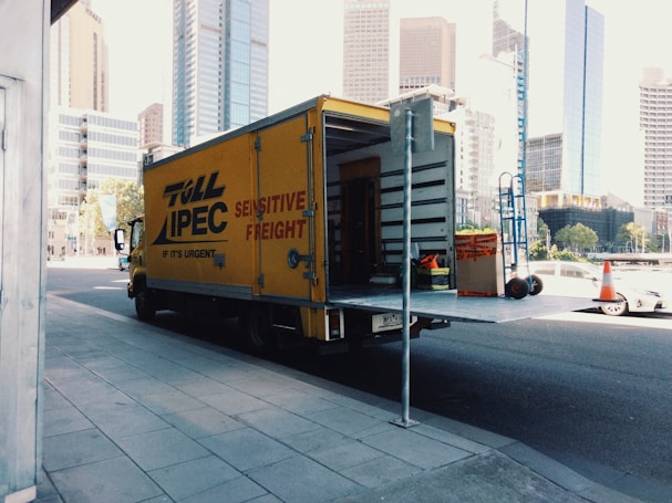 A yellow delivery truck parked on a city street with its rear door open, revealing some equipment and boxes inside. The truck has 'Toll IPEC Sensitive Freight' written on its side. There are tall modern buildings in the background, and a few traffic cones visible near the sidewalk.