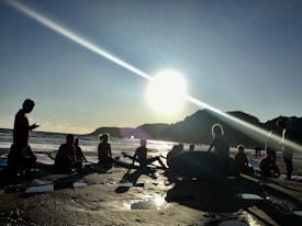 A group of people, silhouetted against a bright, low sun, are gathered on a beach with surfboards as the ocean waves are visible in the background. The scene conveys a calm and communal atmosphere, with reflections of light on the wet sand creating an intriguing play of shadows.