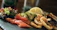 A dish featuring a variety of foods is elegantly arranged on a dark slate surface. The arrangement includes fresh green leafy lettuce, thinly sliced salmon, cucumber slices, golden fried calamari, and small fried fish. A lemon wedge provides a hint of citrus freshness.