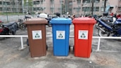 There are three recycling bins placed side by side in an outdoor parking area. Each bin has a different color: brown, blue, and red. The brown bin is labeled for glass only, the blue bin is for paper, and the red bin is for metal and plastic. Behind the bins, several motorcycles are parked in a row, and there is a chain-link fence separating the area from nearby buildings.