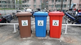 There are three recycling bins placed side by side in an outdoor parking area. Each bin has a different color: brown, blue, and red. The brown bin is labeled for glass only, the blue bin is for paper, and the red bin is for metal and plastic. Behind the bins, several motorcycles are parked in a row, and there is a chain-link fence separating the area from nearby buildings.