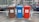 There are three recycling bins placed side by side in an outdoor parking area. Each bin has a different color: brown, blue, and red. The brown bin is labeled for glass only, the blue bin is for paper, and the red bin is for metal and plastic. Behind the bins, several motorcycles are parked in a row, and there is a chain-link fence separating the area from nearby buildings.