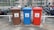 There are three recycling bins placed side by side in an outdoor parking area. Each bin has a different color: brown, blue, and red. The brown bin is labeled for glass only, the blue bin is for paper, and the red bin is for metal and plastic. Behind the bins, several motorcycles are parked in a row, and there is a chain-link fence separating the area from nearby buildings.