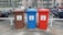 There are three recycling bins placed side by side in an outdoor parking area. Each bin has a different color: brown, blue, and red. The brown bin is labeled for glass only, the blue bin is for paper, and the red bin is for metal and plastic. Behind the bins, several motorcycles are parked in a row, and there is a chain-link fence separating the area from nearby buildings.