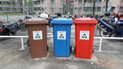 There are three recycling bins placed side by side in an outdoor parking area. Each bin has a different color: brown, blue, and red. The brown bin is labeled for glass only, the blue bin is for paper, and the red bin is for metal and plastic. Behind the bins, several motorcycles are parked in a row, and there is a chain-link fence separating the area from nearby buildings.