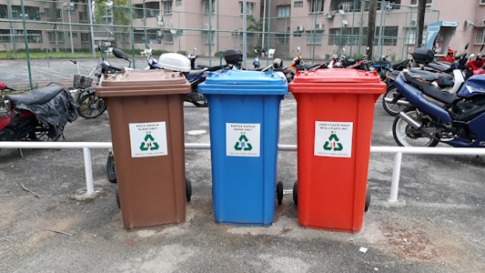 There are three recycling bins placed side by side in an outdoor parking area. Each bin has a different color: brown, blue, and red. The brown bin is labeled for glass only, the blue bin is for paper, and the red bin is for metal and plastic. Behind the bins, several motorcycles are parked in a row, and there is a chain-link fence separating the area from nearby buildings.