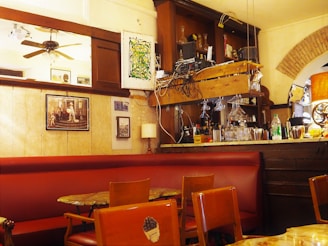 A cozy bar interior featuring a variety of furniture and decor. Wooden chairs and tables with a red cushioned bench line the seating area. The wall has framed artworks and a large mirror. A wooden bar counter is topped with glasses, bottles, and overhead glass racks. A mix of rustic wood and modern elements give the space a quaint charm.