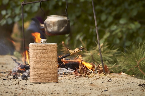 A rustic leather-wrapped flask placed beside a campfire, with a blurred background of trees and evening sky.