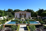 A beautifully manicured garden features rectangular pools surrounded by lush green hedges and potted plants. At the center, a classical statue and fountain add elegance to the symmetrical design. A classical building with arches and a tiled roof stands in the background, framed by dense trees and shrubs.