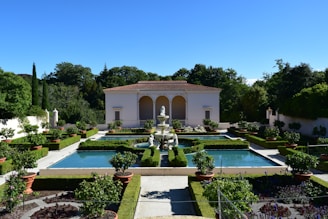 A beautifully manicured garden features rectangular pools surrounded by lush green hedges and potted plants. At the center, a classical statue and fountain add elegance to the symmetrical design. A classical building with arches and a tiled roof stands in the background, framed by dense trees and shrubs.