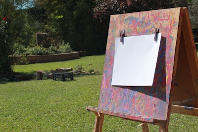 An easel stands in a grassy outdoor setting, holding a blank white canvas. The easel is splattered with colorful paint, suggesting frequent artistic use. In the background, there is lush greenery and a garden bed, creating a serene and natural environment.
