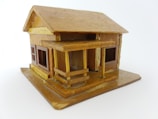 A small, intricately crafted wooden model house with a pitched roof and a porch. The structure includes windows with visible frames, and the overall design appears detailed, reflecting careful craftsmanship.