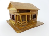 A small, intricately crafted wooden model house with a pitched roof and a porch. The structure includes windows with visible frames, and the overall design appears detailed, reflecting careful craftsmanship.