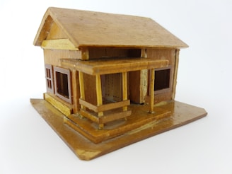 A small, intricately crafted wooden model house with a pitched roof and a porch. The structure includes windows with visible frames, and the overall design appears detailed, reflecting careful craftsmanship.
