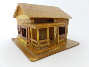 A small, intricately crafted wooden model house with a pitched roof and a porch. The structure includes windows with visible frames, and the overall design appears detailed, reflecting careful craftsmanship.