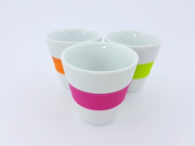 Three white ceramic cups with colorful silicone bands around the middle. The bands are in pink, orange, and green, and each cup is uniformly shaped with a smooth finish.