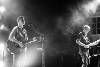 Two musicians are performing on stage, both playing guitars and surrounded by stage lighting and smoke effects. One musician is singing into a microphone while playing an acoustic guitar, while the other plays an electric guitar.