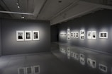 A modern art gallery interior with gray walls and polished floors. Several framed black-and-white photographs are displayed on two walls, each illuminated by spotlights. The room is dimly lit, creating a contemplative atmosphere.