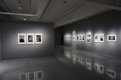 A modern art gallery interior with gray walls and polished floors. Several framed black-and-white photographs are displayed on two walls, each illuminated by spotlights. The room is dimly lit, creating a contemplative atmosphere.