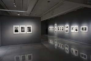 A modern art gallery interior with gray walls and polished floors. Several framed black-and-white photographs are displayed on two walls, each illuminated by spotlights. The room is dimly lit, creating a contemplative atmosphere.