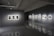 A modern art gallery interior with gray walls and polished floors. Several framed black-and-white photographs are displayed on two walls, each illuminated by spotlights. The room is dimly lit, creating a contemplative atmosphere.