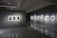 A modern art gallery interior with gray walls and polished floors. Several framed black-and-white photographs are displayed on two walls, each illuminated by spotlights. The room is dimly lit, creating a contemplative atmosphere.