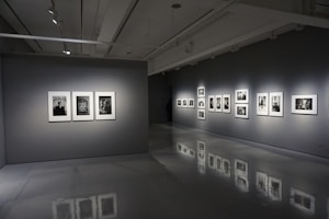 A modern art gallery interior with gray walls and polished floors. Several framed black-and-white photographs are displayed on two walls, each illuminated by spotlights. The room is dimly lit, creating a contemplative atmosphere.