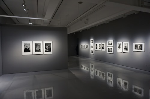 A modern art gallery interior with gray walls and polished floors. Several framed black-and-white photographs are displayed on two walls, each illuminated by spotlights. The room is dimly lit, creating a contemplative atmosphere.