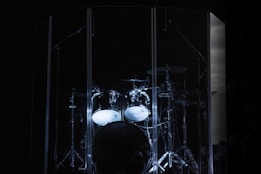 A drum kit is set within a transparent acoustic shield on a dark stage. The drums are partially illuminated, emphasizing their metallic hardware and glossy surfaces.