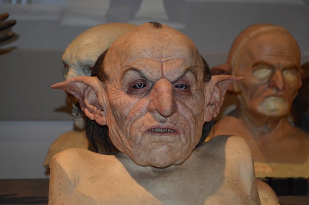 A detailed, lifelike mask of a grotesque fantasy creature with pointed ears and prominent facial features is prominently displayed. The mask appears to be part of a collection, with similar masks in the background showcasing varying expressions and styles.