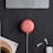 A red smart speaker is placed on a dark surface alongside a partial view of a laptop, white wireless earbuds, and their charging case. A portion of a white object with a cork surface is also visible.